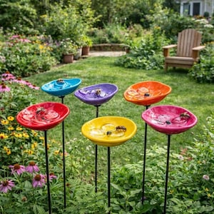Puede incluir: Seis coloridos bebederos de cerámica para pájaros en un jardín. Cada uno es de un color diferente: azul, morado, naranja, rojo, amarillo y rosa. Montados sobre estacas de metal negro, atraen insectos.