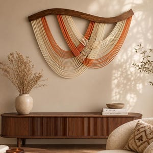Puede incluir: Un tapiz de pared con un marco de madera oscura curvada y múltiples hilos de cuerda color crema y naranja quemado dispuestos en un patrón superpuesto. La cuerda forma una serie de curvas profundas suspendidas del arco de madera.