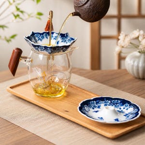 Könnte beinhalten: Eine klare Glas-Teekanne mit Holzgriff und floralem Design steht auf einem Holztablett. Ein blau-weißes Blumensieb befindet sich auf der Teekanne. Eine braune Teekanne gießt Tee in das Sieb. Eine passende Untertasse ist ebenfalls auf dem Tablett.