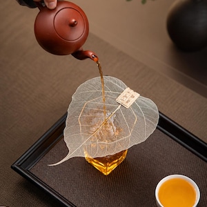 May include: A brown teapot pours tea over a leaf-shaped tea bag into a glass. The tea is amber-colored, and the scene is set on a black tray with small white teacups. The background is a neutral brown.