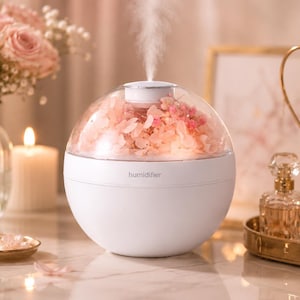 May include: A spherical white humidifier with a clear dome top containing pink dried flowers. The device emits a fine mist from the centre and features the word humidifier printed in small grey letters on the front of the smooth white base.