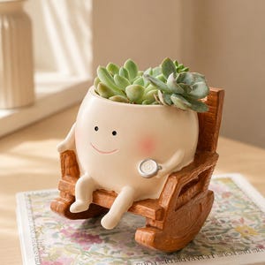 May include: A whimsical, cream-colored planter shaped like a smiling character, complete with rosy cheeks, holding a stethoscope, and seated in a miniature wooden rocking chair. The planter is filled with green succulents. The base of the planter is resting on a book.