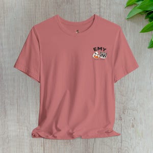 Może przedstawiać: T-shirt z krótkim rękawem w kolorze przybrudzonego różu z małą grafiką na lewej piersi. Grafika przedstawia napis EMY nad rysunkowymi ilustracjami kaczki i szopa pracza, z mniejszym napisem DEMY REMY wydrukowanym pod postaciami.