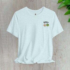May include: A light blue short-sleeve t-shirt with a crew neckline. On the upper left chest, there is a small graphic featuring the text EMY above a cartoon white duck and a green frog character, with the smaller text DEMY-FEMY below them.