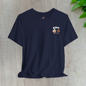 May include: A navy blue short-sleeve t-shirt featuring a small graphic on the upper left chest. The graphic displays the white text EMY above two cartoon character heads, one white and one brown, with the text DEMY PEMY printed below them.