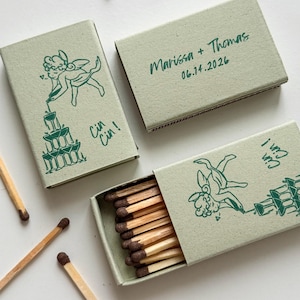 May include: Three light green matchboxes with a green illustration of a cherub pouring champagne into a tower of glasses. One box is open, revealing the matches. The text "Cin Cin!" is printed on the boxes. The top of one box reads "Marissa + Thomas 06.14.2026".