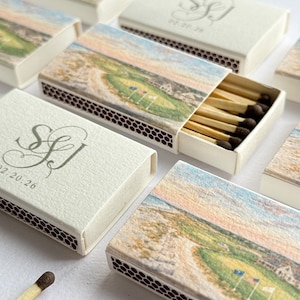 May include: Ivory matchboxes with a golf course landscape design. The boxes feature a printed landscape scene with a green golf course, blue flags, and a coastal view. Some boxes have initials and a date printed on them. The matches are visible inside an open box.