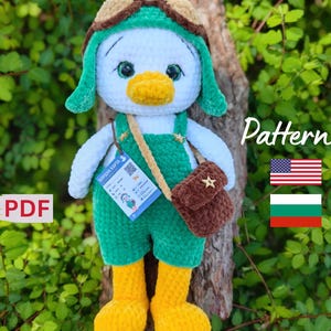May include: A handmade crochet duck doll wearing a green aviator hat and overalls, with yellow boots and a brown satchel. The doll has a white body, a yellow beak, and green eyes. A tag is attached to the doll. The word "Pattern" is visible.