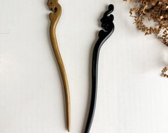 May include: Two hair sticks, one light brown and one black, each featuring a carved, stylized animal head at the top. The hair sticks have a smooth, curved design and are positioned vertically against a plain white background.