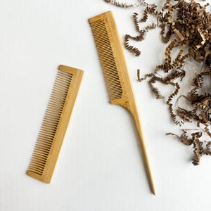 May include: Two bamboo combs on a white surface. One comb has a handle, and the other is a smaller, rectangular comb. The combs are light brown with thin, evenly spaced teeth. Brown paper shreds are scattered nearby.