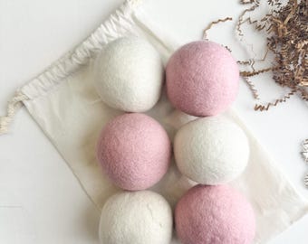 May include: Six spherical wool dryer balls arranged in two columns on a cream-colored cotton drawstring bag. Three balls are solid white and three are solid dusty pink. The texture of the wool is felted and matte, showing a soft, rounded surface.