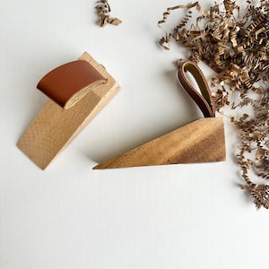 Op de afbeelding: Twee houten deurvastzetters met bruine leren details. Eén heeft een leren lus, de andere een riem. De wigvormige deurvastzetters zijn gemaakt van lichtgekleurd hout en houden deuren open.