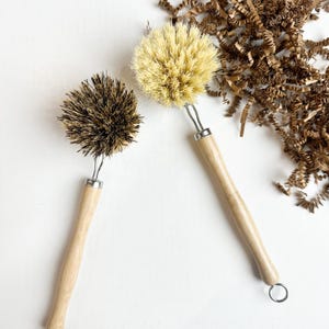 Può includere: Due spazzole per piatti con manici in legno e setole naturali. Una spazzola ha setole marrone chiaro, l'altra ha setole marrone scuro. Le spazzole hanno un anello di metallo per appenderle. Sullo sfondo ci sono trucioli di carta marrone.