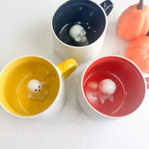 May include: Three ceramic mugs with white exteriors and colored interiors. One mug has a black interior with a white skull at the bottom, one has a yellow interior with a white ghost, and one has a red interior with a white figure with red eyes.