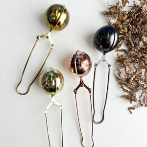 Puede incluir: Cuatro infusores de té metálicos en dorado, oro rosa, negro y latón. Cada infusor tiene una esfera perforada para hojas de té, unida a un asa con un mecanismo de presión. Los infusores están dispuestos sobre una superficie blanca.