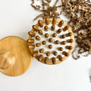 Peut inclure: Un outil de massage en bois avec une tête ronde et texturée et une poignée séparée. La tête comporte de nombreux petits chevilles en bois arrondies sur une surface blanche. La poignée et la base sont en bambou clair.