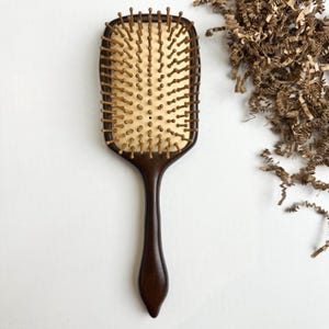 Puede incluir: Un cepillo de pelo de madera marrón oscuro con una cabeza rectangular y un mango largo. El cepillo tiene cerdas de color marrón claro y un cojín de color beige claro. Una herramienta clásica para el cuidado del cabello.