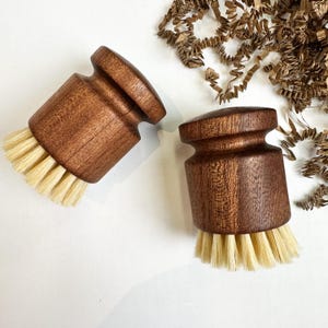 Puede incluir: Dos cepillos de madera para lavar platos con cerdas de color claro. Los cepillos tienen un dise&ntilde;o redondeado y ergon&oacute;mico y est&aacute;n hechos de madera marr&oacute;n oscura. Las cerdas est&aacute;n muy juntas y parecen estar hechas de fibras naturales. El fondo es blanco con papel marr&oacute;n.