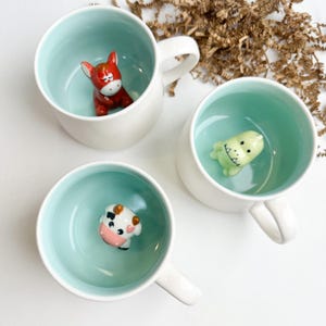 May include: Three white ceramic mugs with light blue interiors, each featuring a small, three-dimensional animal figure at the bottom. The figures include a red donkey, a light green dinosaur, and a black and white cow with pink accents.