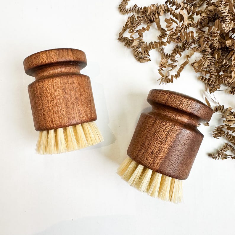 Puede incluir: Dos cepillos faciales de madera con cerdas de color claro. Los cepillos tienen un cuerpo cil&iacute;ndrico y una parte superior redondeada. La madera tiene un color marr&oacute;n oscuro intenso. Las cerdas son de un color natural y suave. Los cepillos se muestran sobre una superficie blanca.