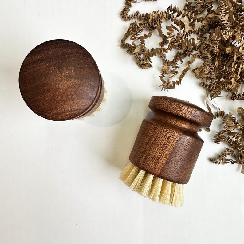 Puede incluir: Dos cepillos de madera para fregar platos con cerdas de color claro. Los cepillos tienen un mango de madera circular de color marr&oacute;n oscuro. Un cepillo est&aacute; en posici&oacute;n vertical y el otro est&aacute; acostado de lado. Tiras de papel marr&oacute;n en el fondo sobre una superficie blanca.