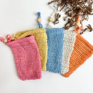 May include: Five colorful soap saver bags in pink, yellow, blue, white, and orange. Each bag is made of a woven material and has a drawstring closure with wooden beads. The bags are arranged on a white surface.