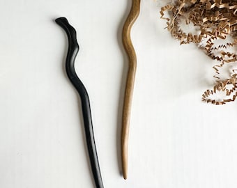 May include: Two hair sticks with a smooth, wavy, and elongated design. One hair stick is finished in a solid matte black, while the other is finished in a natural light brown wood tone. Both items feature a tapered point at the bottom end.