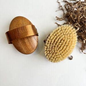Op de afbeelding: Twee houten lichaamsborstels op een witte ondergrond. Eén is ovaal van vorm met een geweven band, de andere is een borstel met natuurlijke borstelharen. De borstels zijn voor droogborstelen en exfoliëren.