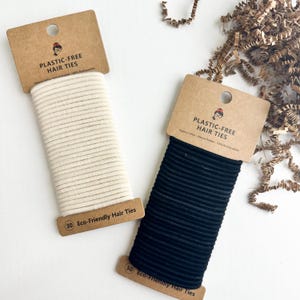 May include: Two spools of hair ties, one cream-colored and one black, are displayed. Each spool is attached to a brown cardboard tag that reads "PLASTIC-FREE HAIR TIES" and "Eco-Friendly Hair Ties". The image is taken from a top-down perspective.