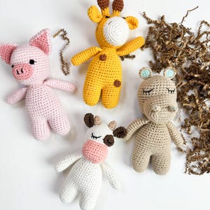 May include: Four handmade crochet animal toys: a pink pig, a yellow giraffe, a white cow, and a tan hippo. Each toy has a unique design, with the giraffe and cow featuring brown spots and the hippo with blue accents.