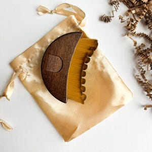 May include: A wooden pottery rib tool with a curved, dark brown handle and a yellow brush head, resting on a gold satin pouch. The tool is designed for shaping and smoothing clay. The pouch is tied with a matching satin ribbon.