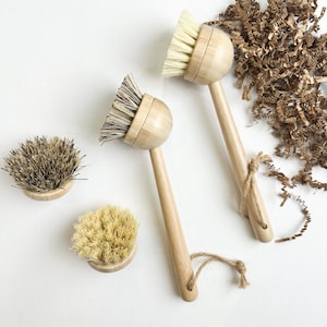 Puede incluir: Dos cepillos de madera para lavar platos con cerdas de color claro y mangos largos, junto con dos cabezales de cepillo de repuesto. Los cepillos tienen un acabado de madera natural y están atados con hilo. Las cerdas son una mezcla de colores claros y oscuros.