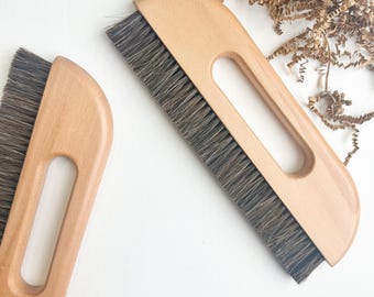 May include: Two rectangular brushes with light-colored wooden handles and dark gray bristles. Each handle features a central elongated cutout for a grip. The brushes are positioned at an angle, showcasing the wood grain and the dense bristle texture.