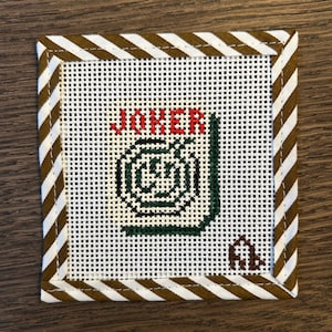 May include: A square needlepoint canvas featuring a white grid background with a brown and white diagonal striped border. The design includes the red text JOKER above a black and green circular graphic and a small brown monogram in the corner.
