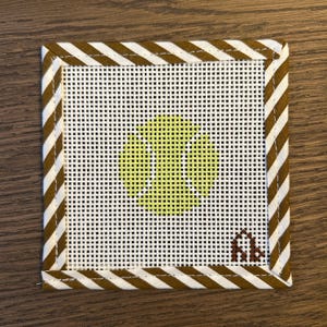 May include: A square needlepoint canvas featuring a white mesh background with a central lime green tennis ball graphic. The border consists of a brown and white diagonal striped fabric binding. A small brown stitched monogram is in the corner.