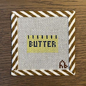 May include: A square needlepoint canvas featuring a white grid background with a yellow rectangular block in the center containing the dark blue text BUTTER. The border consists of a brown and white diagonal striped fabric binding.