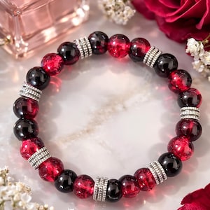 Op de afbeelding: Een armband met afwisselend rode en zwarte ronde kralen, gescheiden door zilverkleurige afstandhouders. De armband wordt getoond met een parfumfles, witte bloemen en een rode roos.