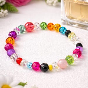 Op de afbeelding: Een kleurrijke kralenarmband met een verscheidenheid aan juweelkleurige kralen. De armband heeft een mix van heldere, roze, groene, oranje, blauwe en zwarte kralen. De armband wordt gepresenteerd op een wit oppervlak met bloemen en een parfumflesje op de achtergrond.