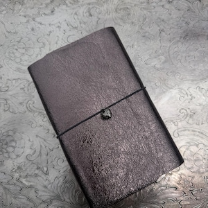 May include: A rectangular dark brown leather journal with a textured surface. The journal is secured by a thin black elastic cord that wraps horizontally around the center, featuring a small, dark metallic heart-shaped charm attached to the cord.