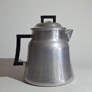 May include: A vintage aluminum coffee pot with a black handle and lid handle. The pot has a spout for pouring and a rounded body. The coffee pot is a silver color and sits on a white surface.