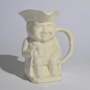 May include: Cream-colored ceramic pitcher in the shape of a seated man wearing a hat and coat. The handle is a curved branch shape. The pitcher has a smiling face and is likely a vintage collectible.