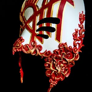 May include: A white half-face mask featuring intricate red and gold filigree scrollwork along the edges. The upper portion displays a bold red and gold geometric symbol with three horizontal black slashes across the centre of the design.