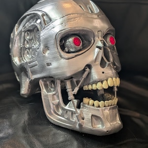 May include: A metallic silver skull with exposed teeth and red glowing eyes. The skull has a detailed mechanical design, with visible gears and robotic components. The jaw is open, and the teeth are a yellowish color.