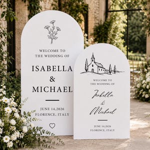 May include: Two white arched wedding signs with black text and illustrations. The taller sign features a floral drawing, the names Isabella & Michael, and the date 14 June 2026. The shorter sign displays a church illustration and the same names.