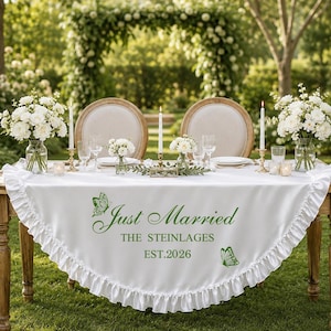 May include: A white ruffled tablecloth featuring green text that reads Just Married THE STEINLAGES EST.2026, accented with two green butterfly illustrations. The cloth is draped over a wooden table set with white flowers, candles, and glassware.