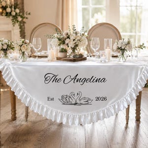 May include: A white satin table runner with a ruffled edge featuring the black printed text The Angelina and the year 2026, accompanied by a graphic illustration of two swans facing each other, centered on the fabric surface.