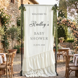 Op de afbeelding: Een hoog, gebroken wit stoffen spandoek opgehangen aan een zwart metalen frame, met de tekst WELCOME TO Radley's BABY SHOWER 15.AUG. 2026. Het spandoek is versierd met olijfgroene linten in de bovenhoeken en heeft een geplooide onderzoom.