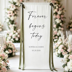 Puede incluir: Un letrero de boda blanco con las palabras "Forever begins Today" en escritura negra. El letrero está adornado con cintas verde oliva y presenta los nombres Sophia & James y la fecha 05.20.2026. El fondo incluye arreglos florales.