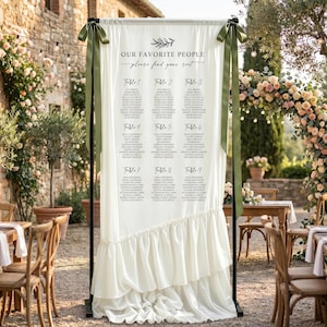 May include: A white fabric wedding seating chart sign suspended on a black metal frame, featuring the text "OUR FAVORITE PEOPLE" and "please find your seat" with nine table lists, accented by olive green ribbons tied at the top corners.