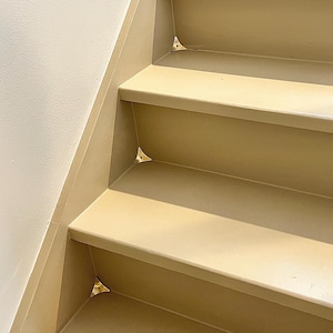 May include: A set of small, triangular, metallic stair corner dust guards installed in the inner corners of beige painted wooden stairs. These polished brass-colored corner protectors are secured with tiny screws at each step junction.
